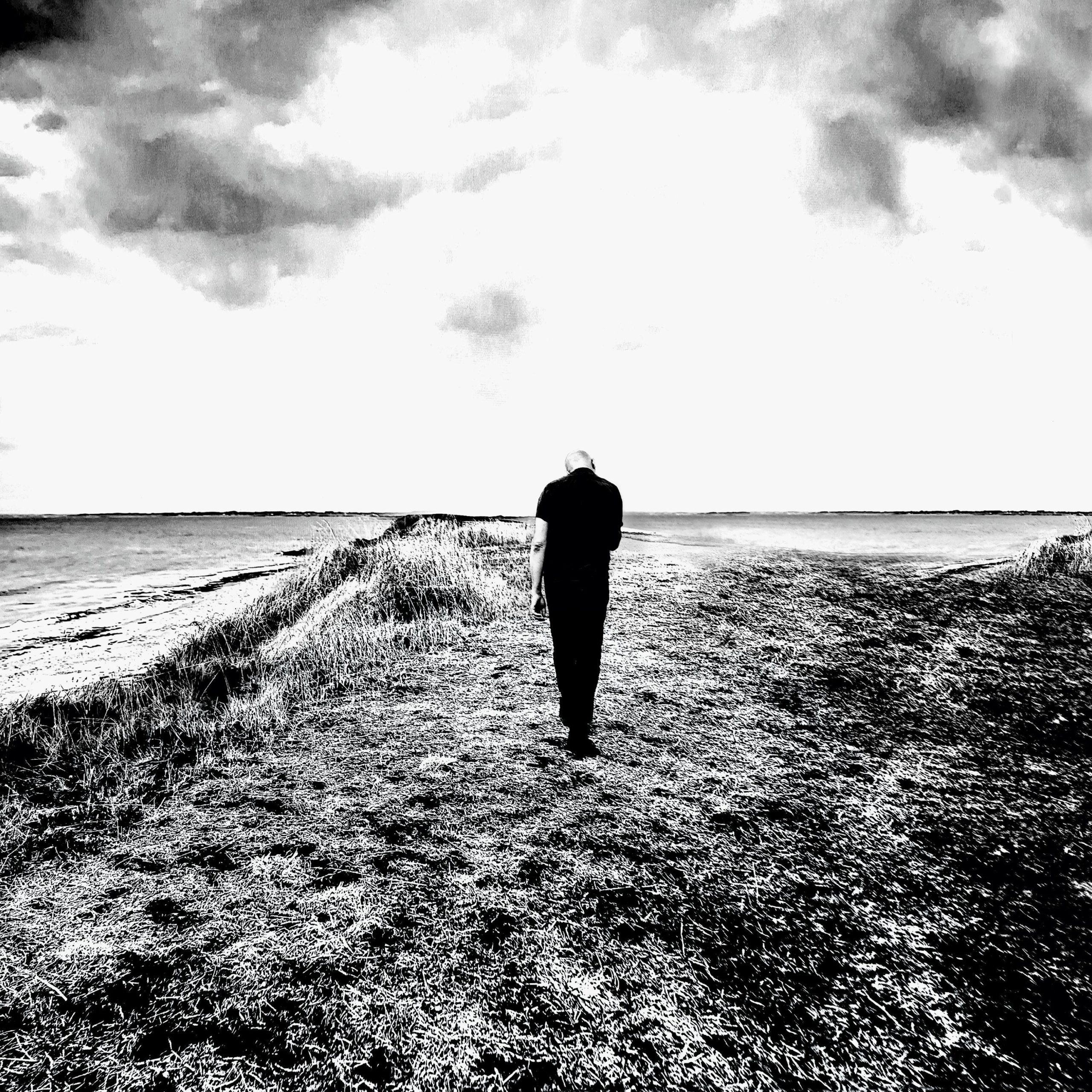 Un paysage en noir et blanc représentant un chemin d'herbe encadré par de l'eau. Un homme de dos marche en direction de l'horizon.